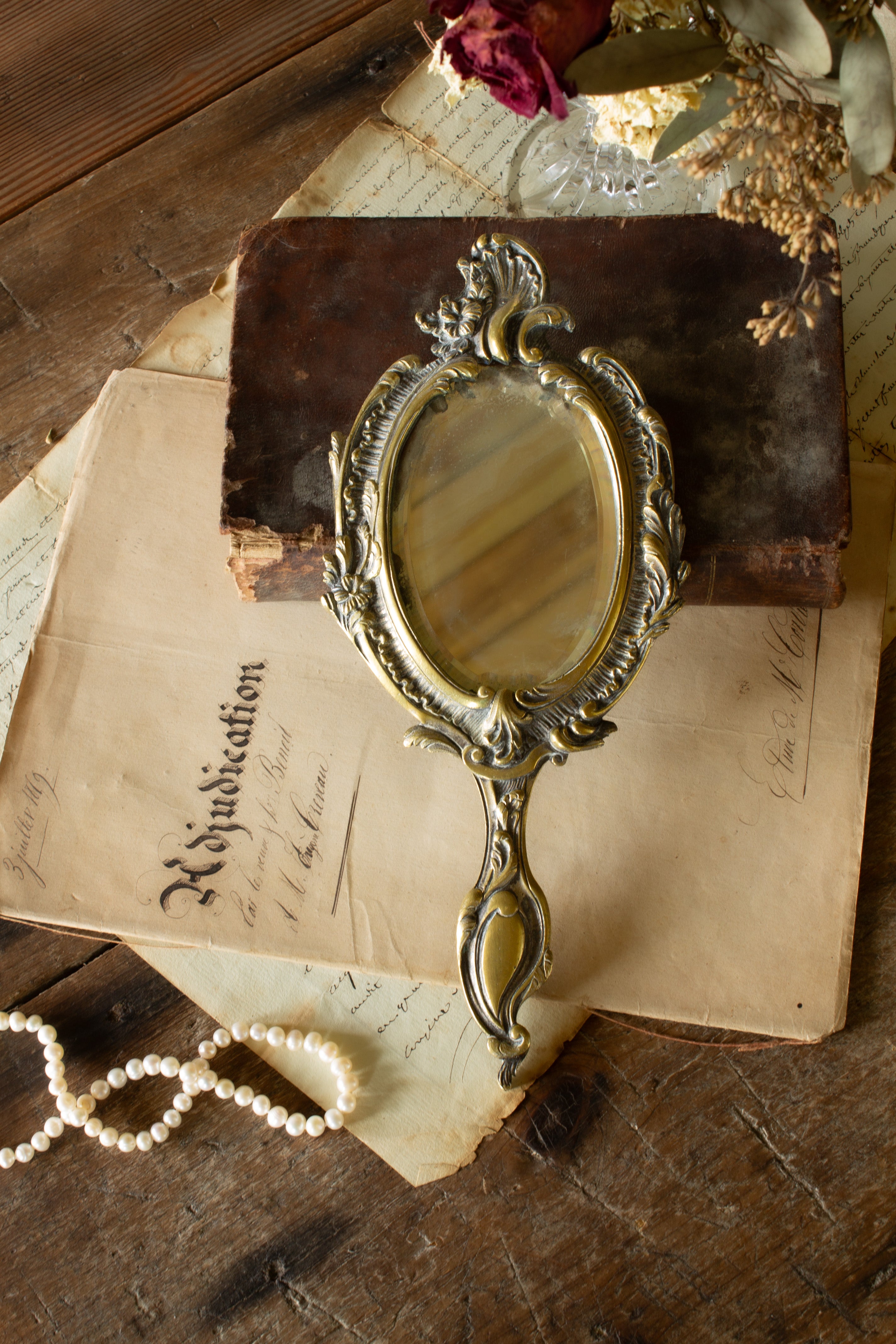 Antique French gilt metal vanity mirror sourced in Paris, with beveled oval glass, Rococo Revival floral and wave-like metalwork, aged bronze-gold finish, handle, and hanging hardware.