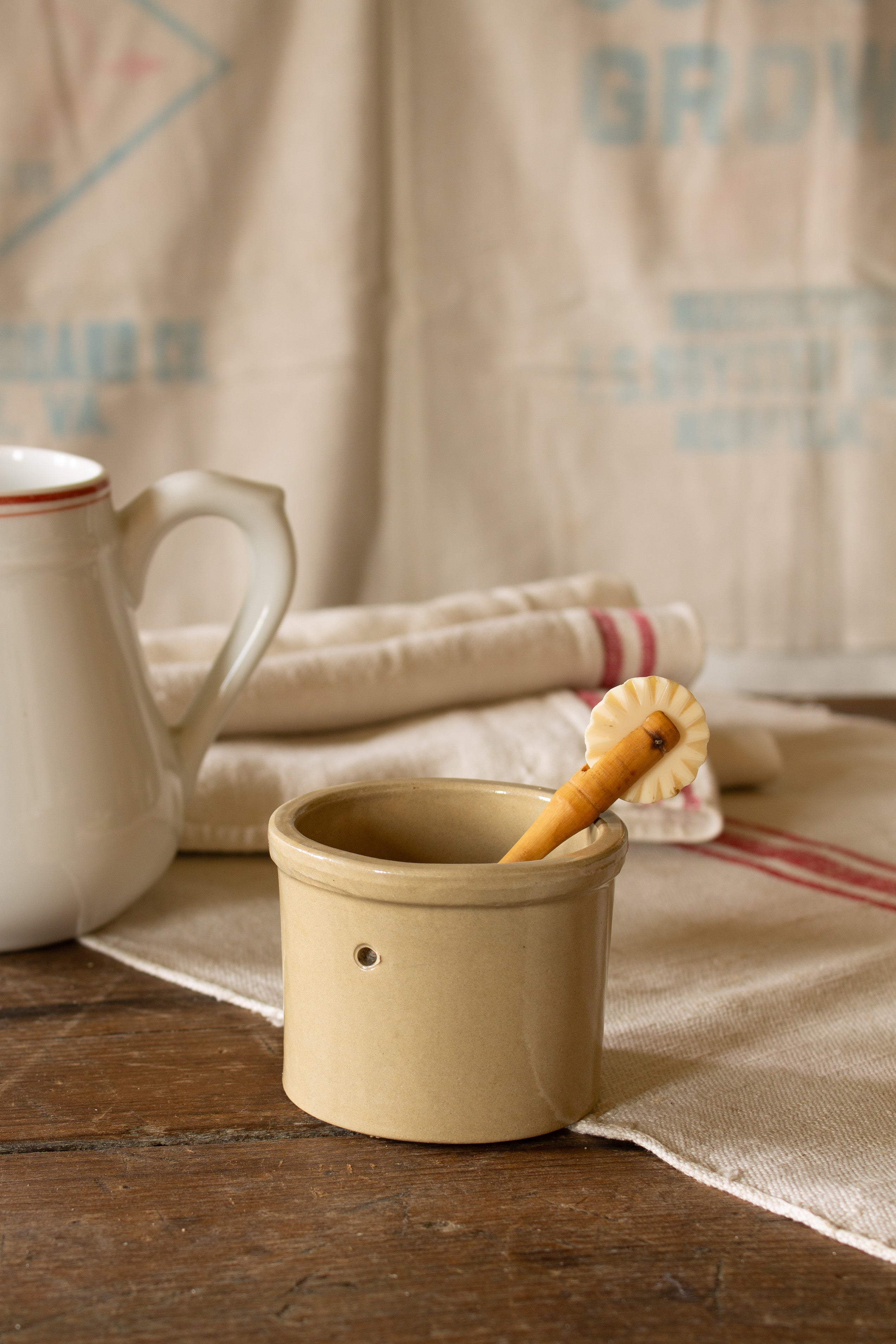 Small vintage unmarked stoneware crock with warm beige brown glaze, thick rolled rim, tiny chip, and circular side indentations, styled with ironstone, linens, and country kitchen pieces.