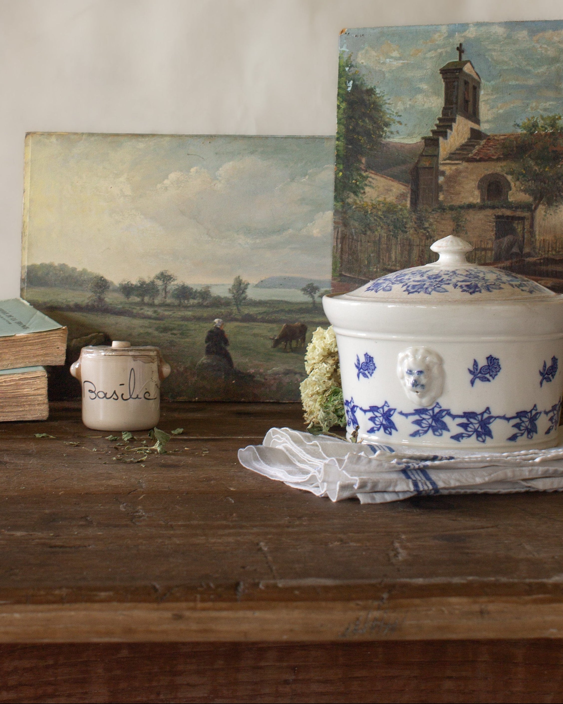 The French Provinces collection featuring antique French landscape paintings, a blue and white tureen, basil crock, old books, and linen on a rustic wood table.