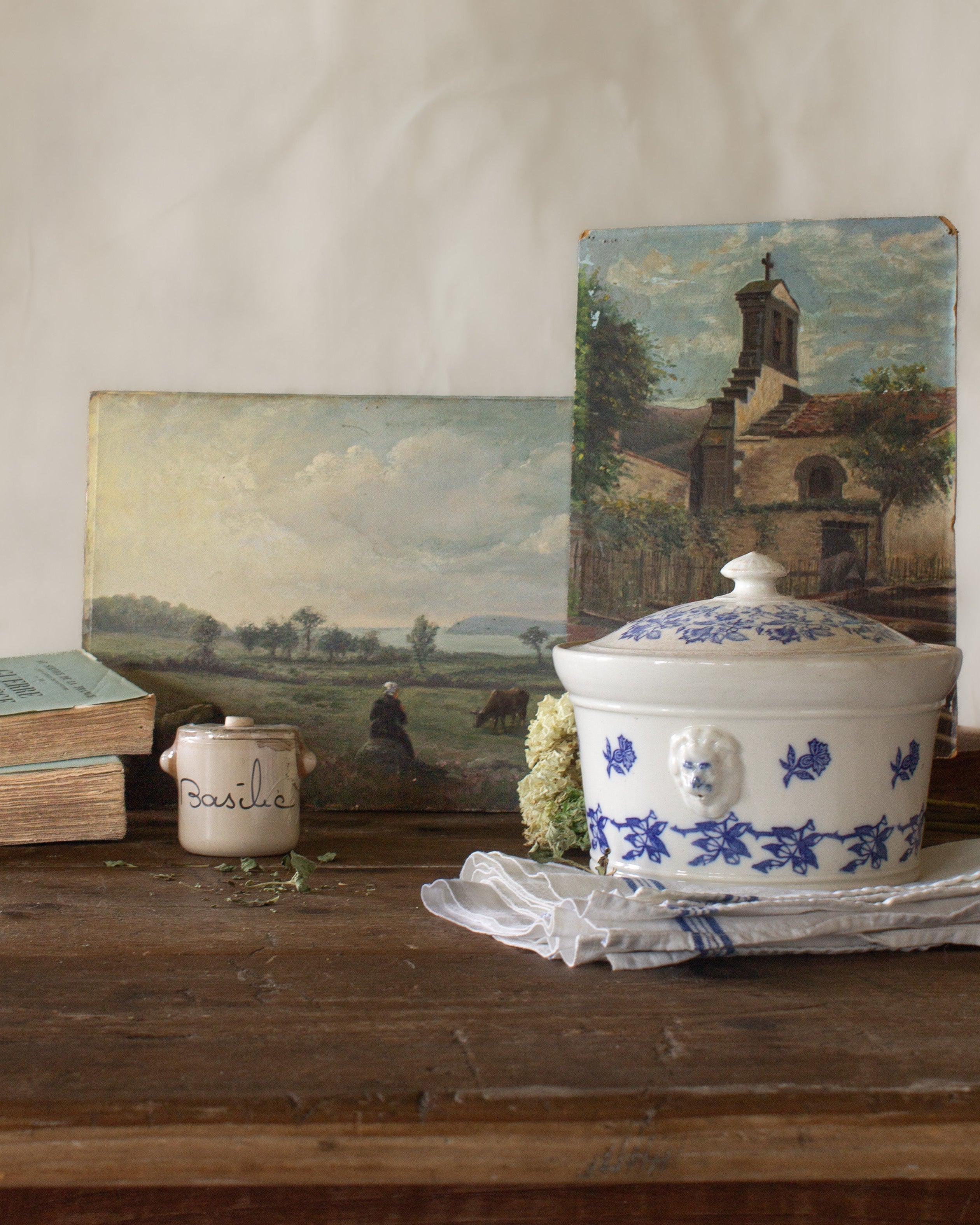 The French Provinces collection featuring antique French landscape paintings, a blue and white tureen, basil crock, old books, and linen on a rustic wood table.