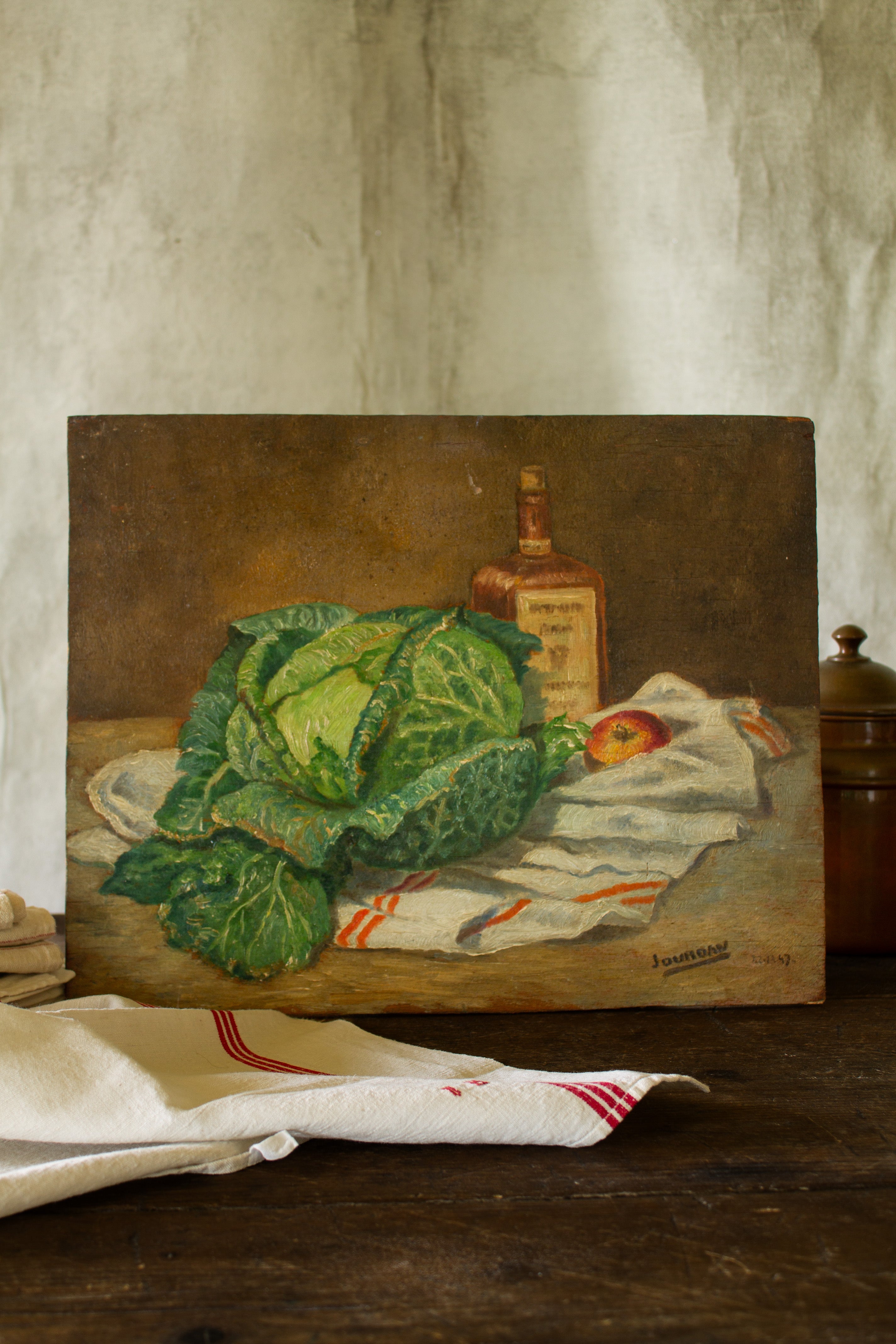 Vintage French 1947 still life painting on wood panel featuring green cabbage, red-striped kitchen torchon, apple, and amber bottle.
