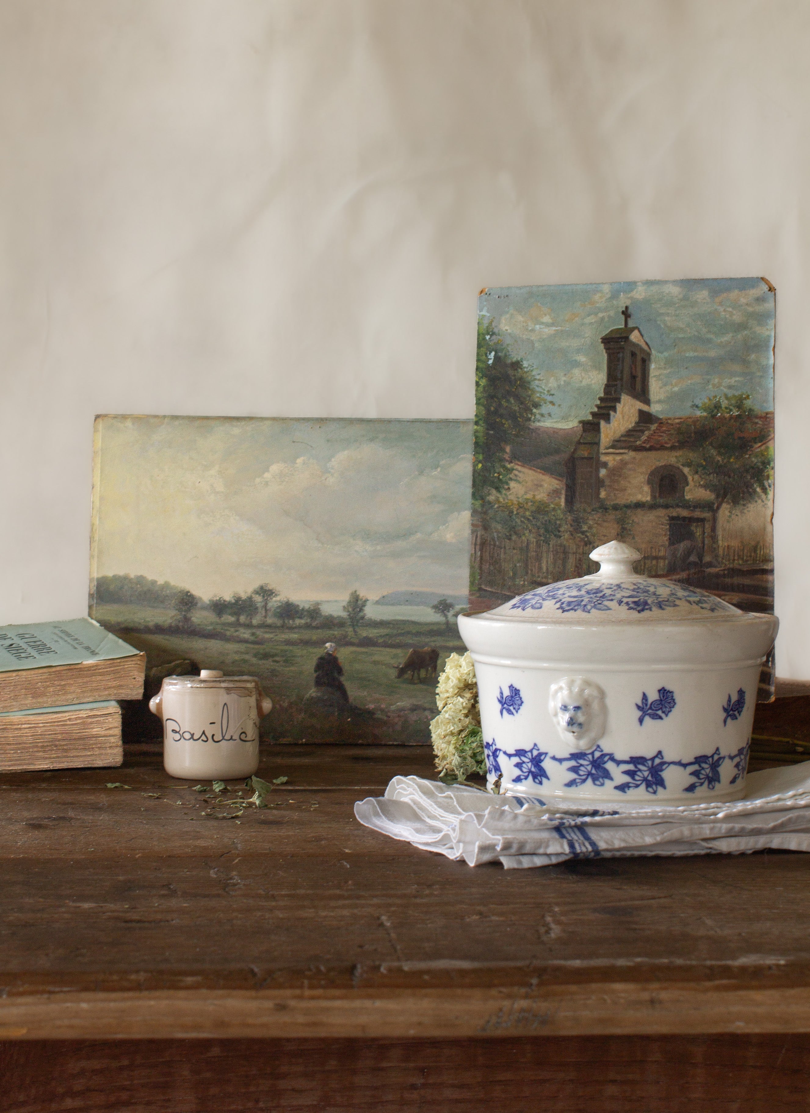 The French Provinces collection featuring antique French landscape paintings, a blue and white tureen, basil crock, old books, and linen on a rustic wood table.