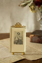 Antique French gilt metal bow-top photo frame sourced in Paris, with ball feet, thick glass, antique photograph of a young girl in a pinafore and knee-high socks, and French photographer’s card with beautiful script lettering on the back.