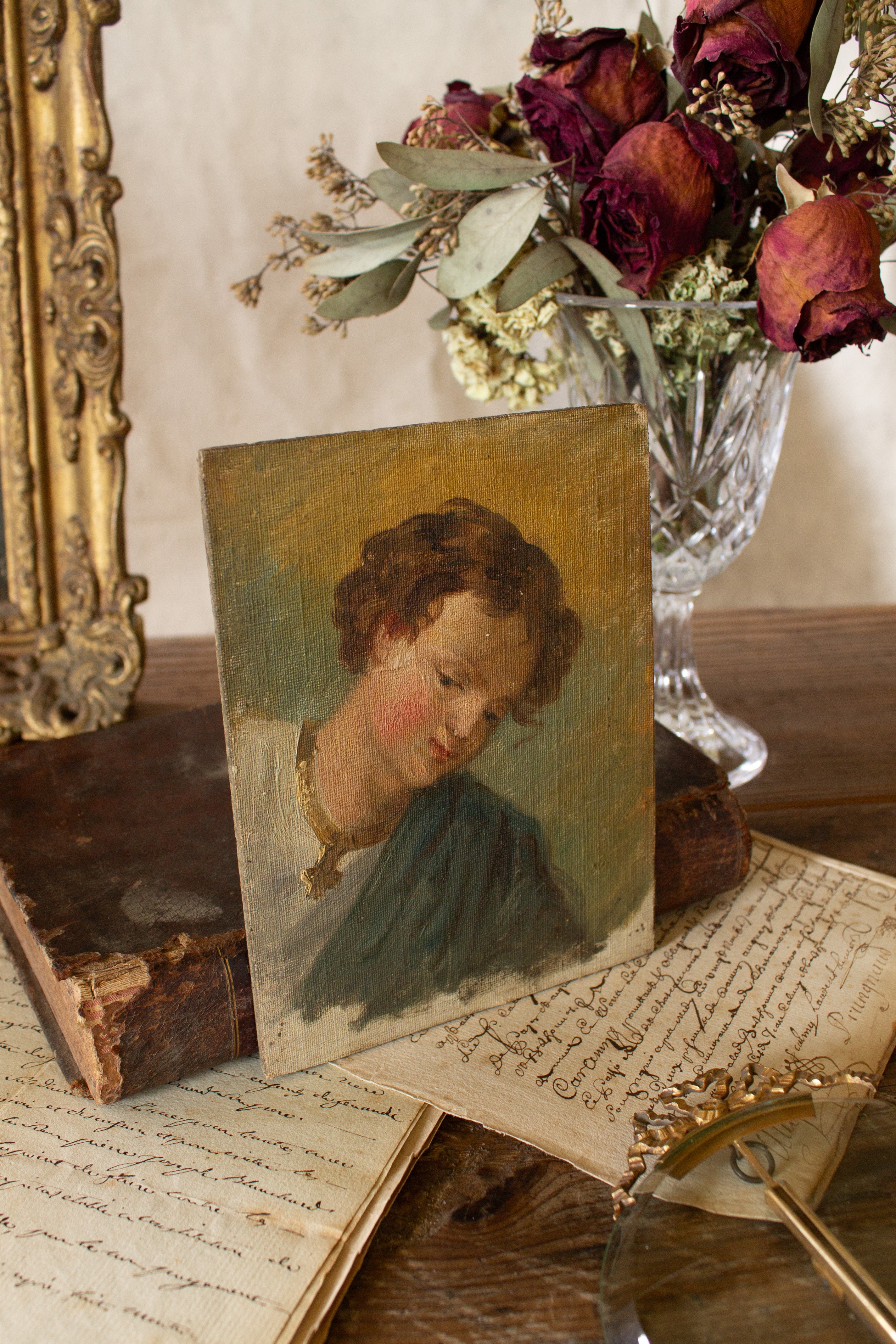 Small antique oil portrait study sourced in Paris, painted on canvas laid to board, showing a young figure with brown curls, flushed cheeks, and a blue-green sash styled with antique books, handwritten French papers, dried roses, and a crystal vase.