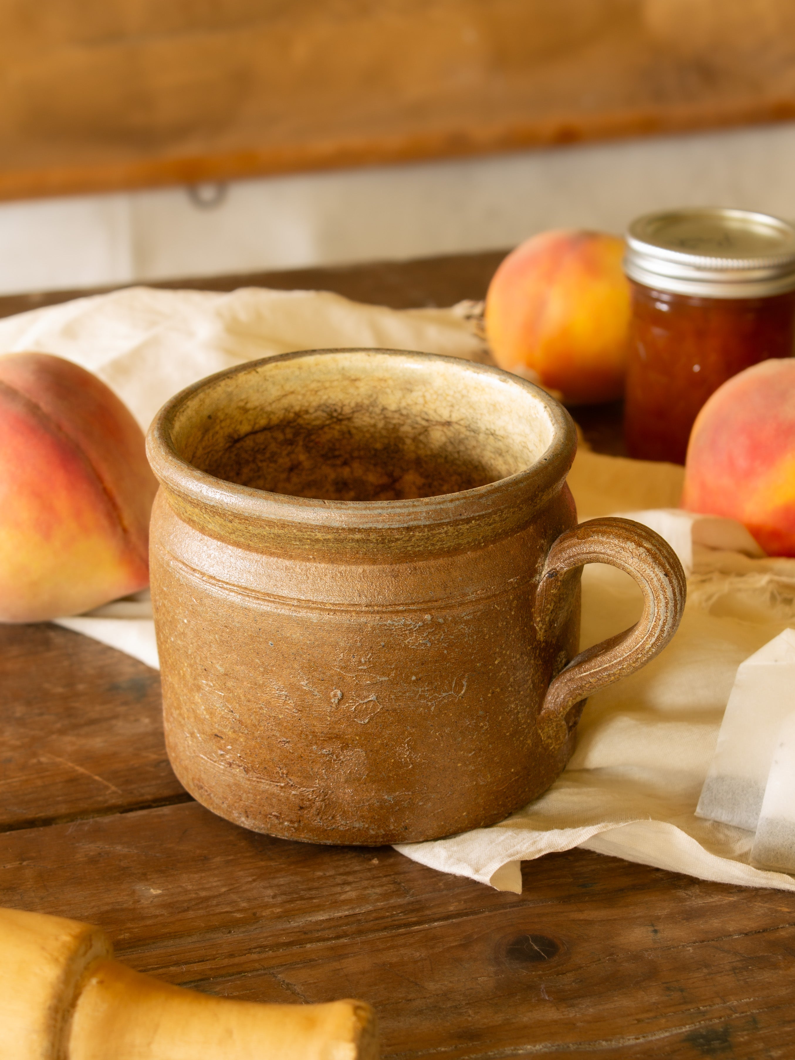 A rustic brown ceramic stoneware mug with a handle, styled on a wooden table with peaches, parchment paper, and a glass jar in the background.