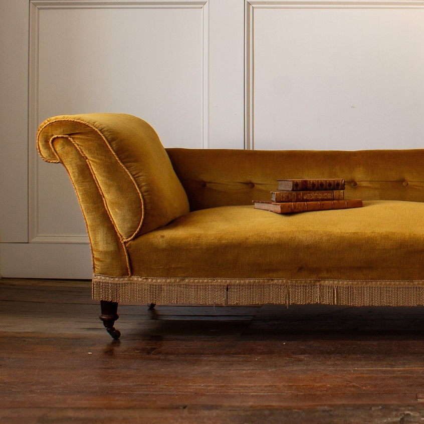 Mustard antique velvet chaise fainting counch in a room with wooden flooring and white paneled walls.