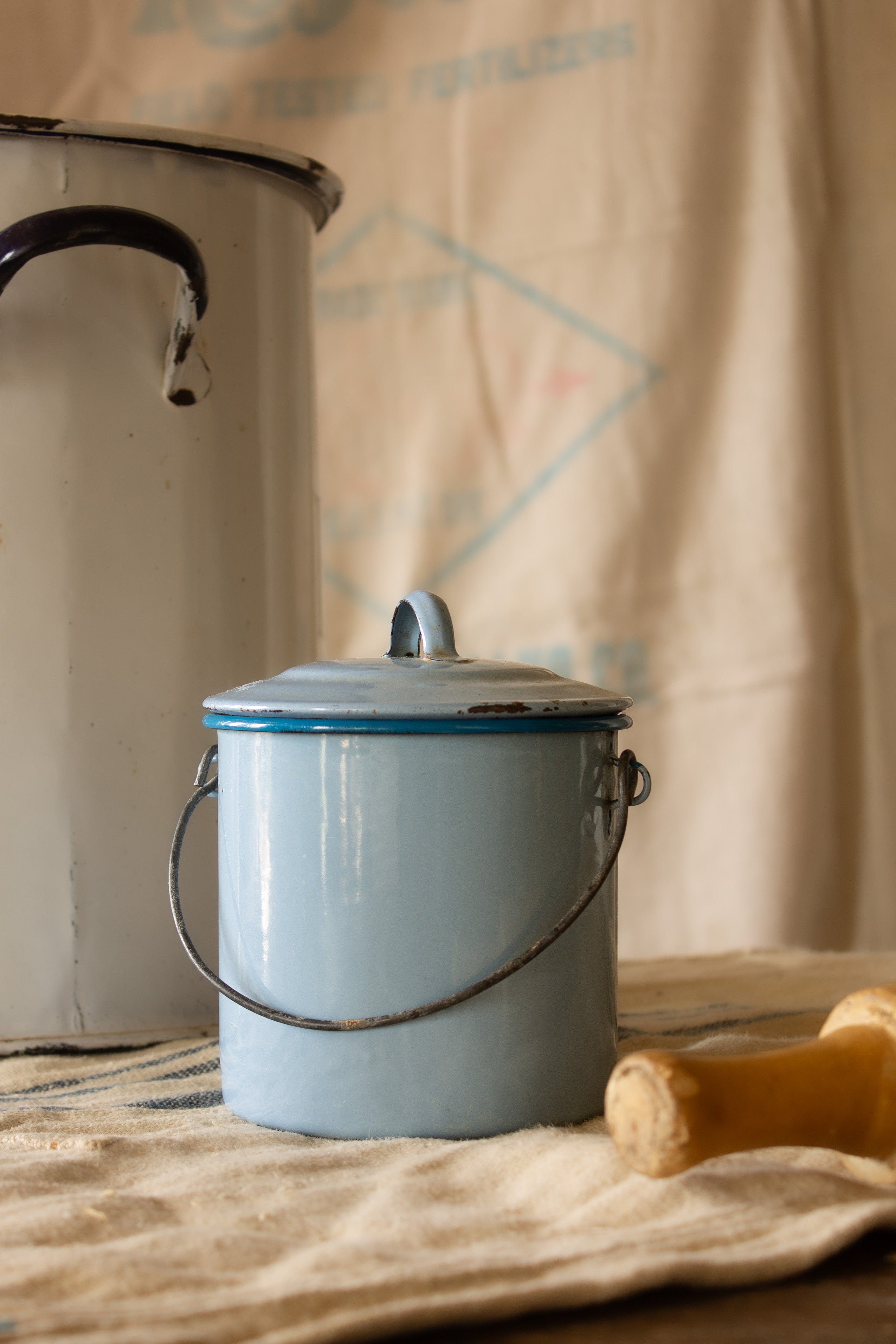 Small French blue enamelware lidded canister with wire handle, worn rim, rust spots, and pale blue enamel surface.