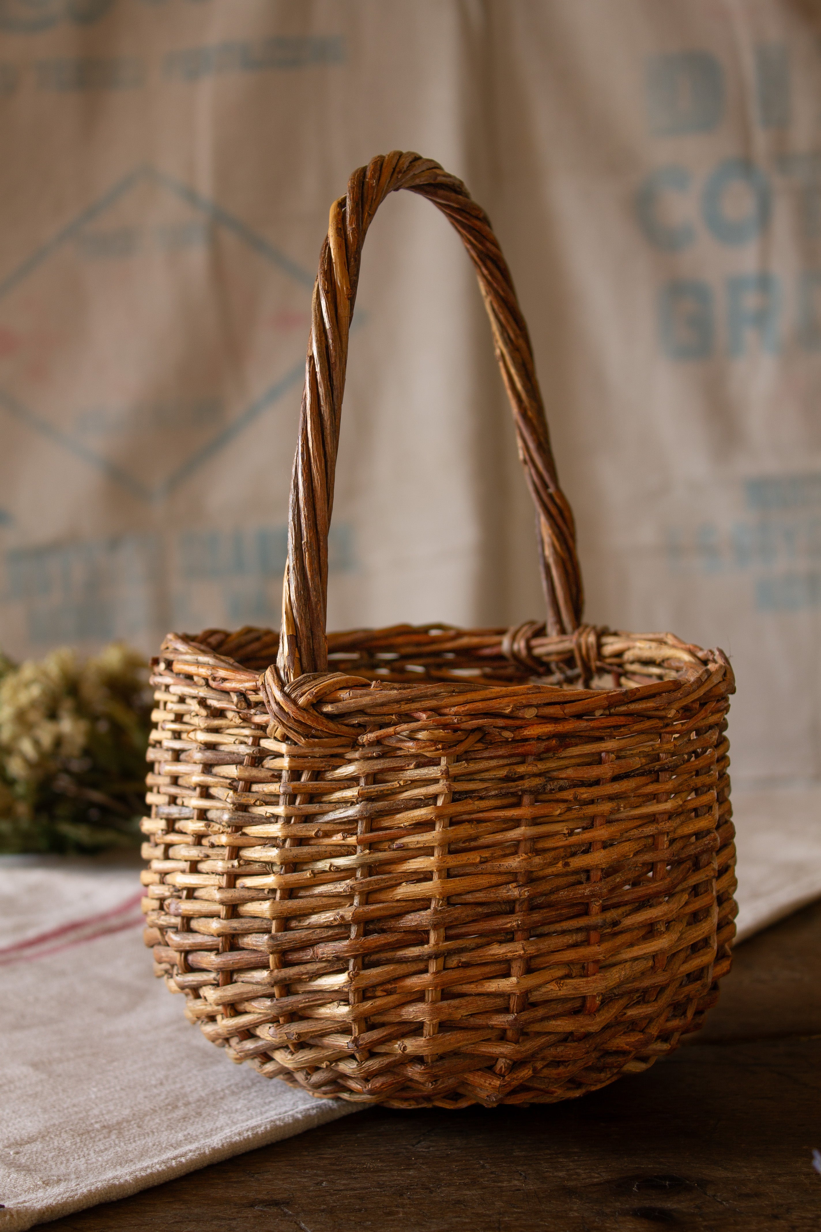 Small handwoven wicker gathering basket with twisted handle and warm rustic patina, styled for primitive country, French country kitchen, farmhouse table, dried flowers, linens, eggs, and seasonal display.