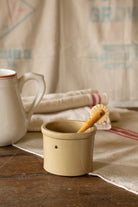 Small vintage unmarked stoneware crock with warm beige brown glaze, thick rolled rim, tiny chip, and circular side indentations, styled with ironstone, linens, and country kitchen pieces.