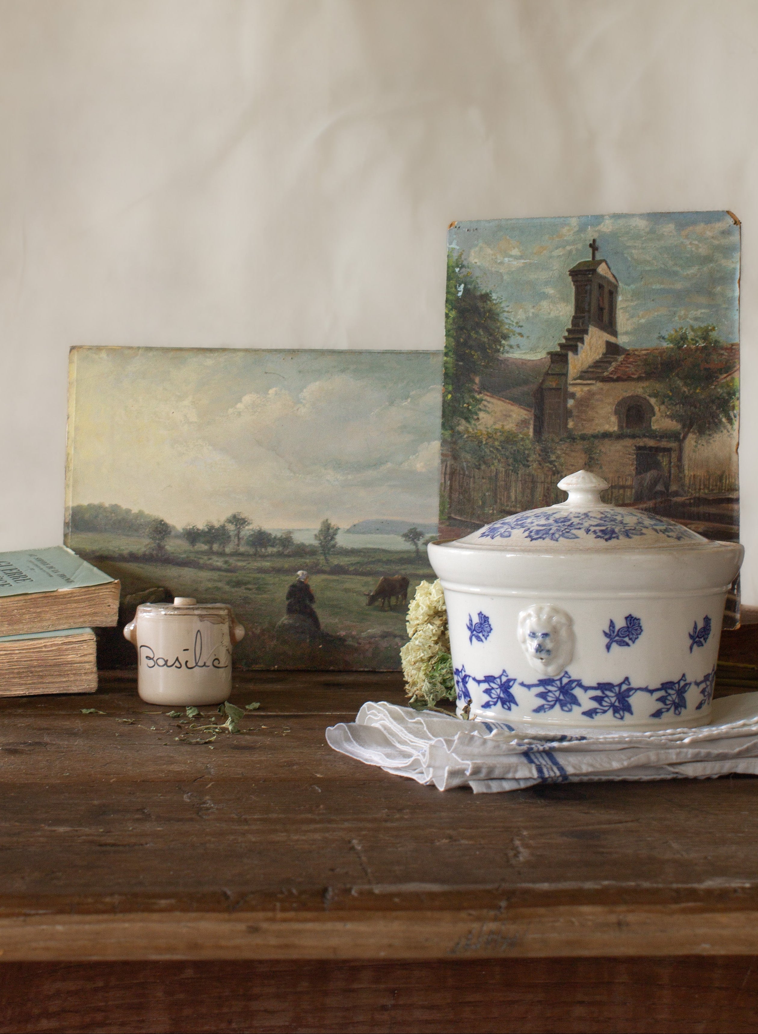 The French Provinces collection featuring antique French landscape paintings, a blue and white tureen, basil crock, old books, and linen on a rustic wood table.