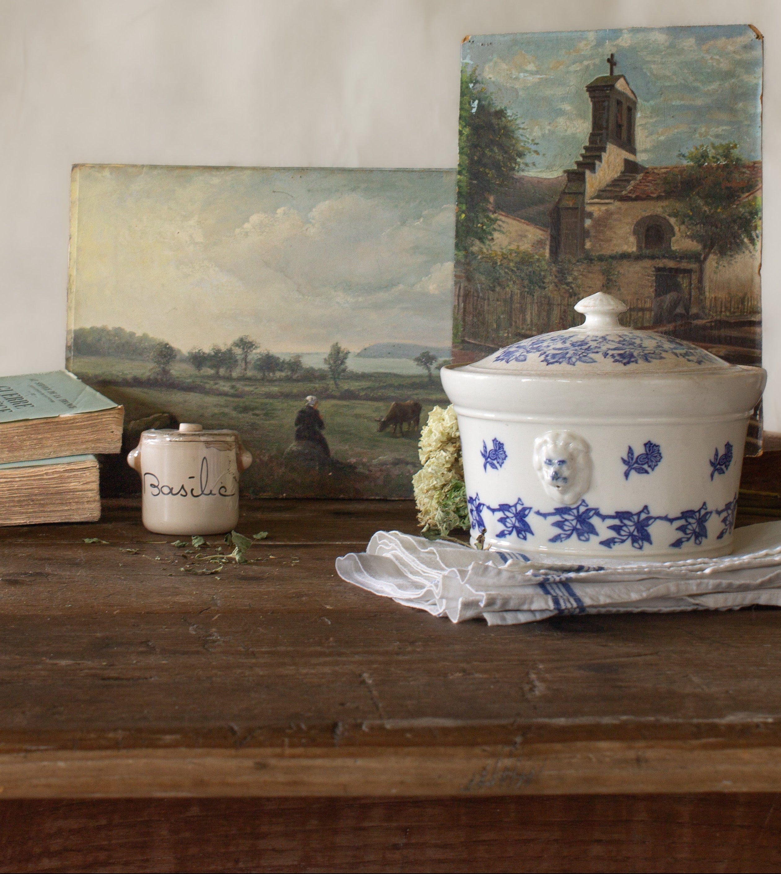 The French Provinces collection featuring antique French landscape paintings, a blue and white tureen, basil crock, old books, and linen on a rustic wood table.
