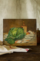 Vintage French 1947 still life painting on wood panel featuring green cabbage, red-striped kitchen torchon, apple, and amber bottle.
