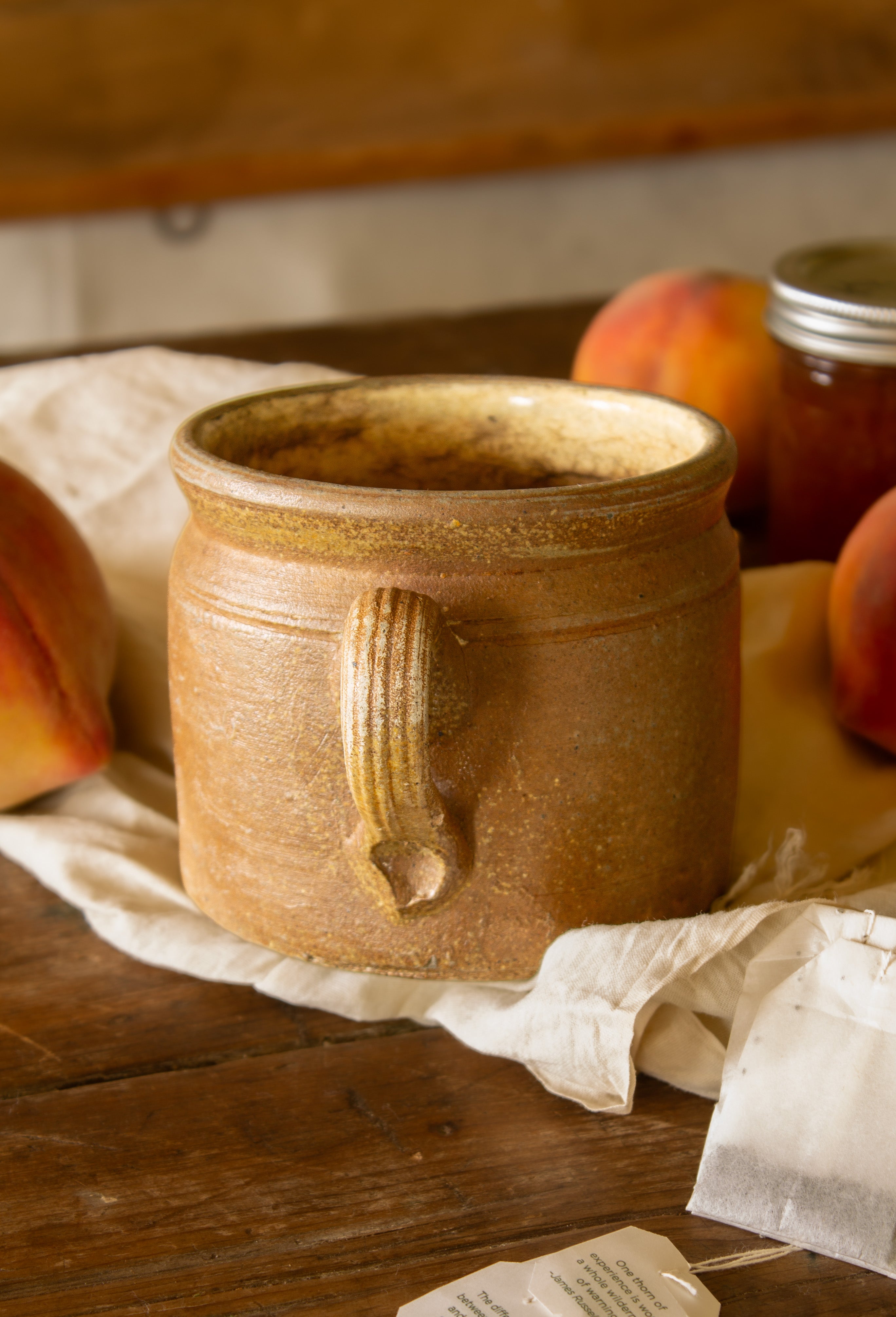 Vintage hand-thrown brown stoneware handled crock displayed with peaches, linen, and a jar of preserves.