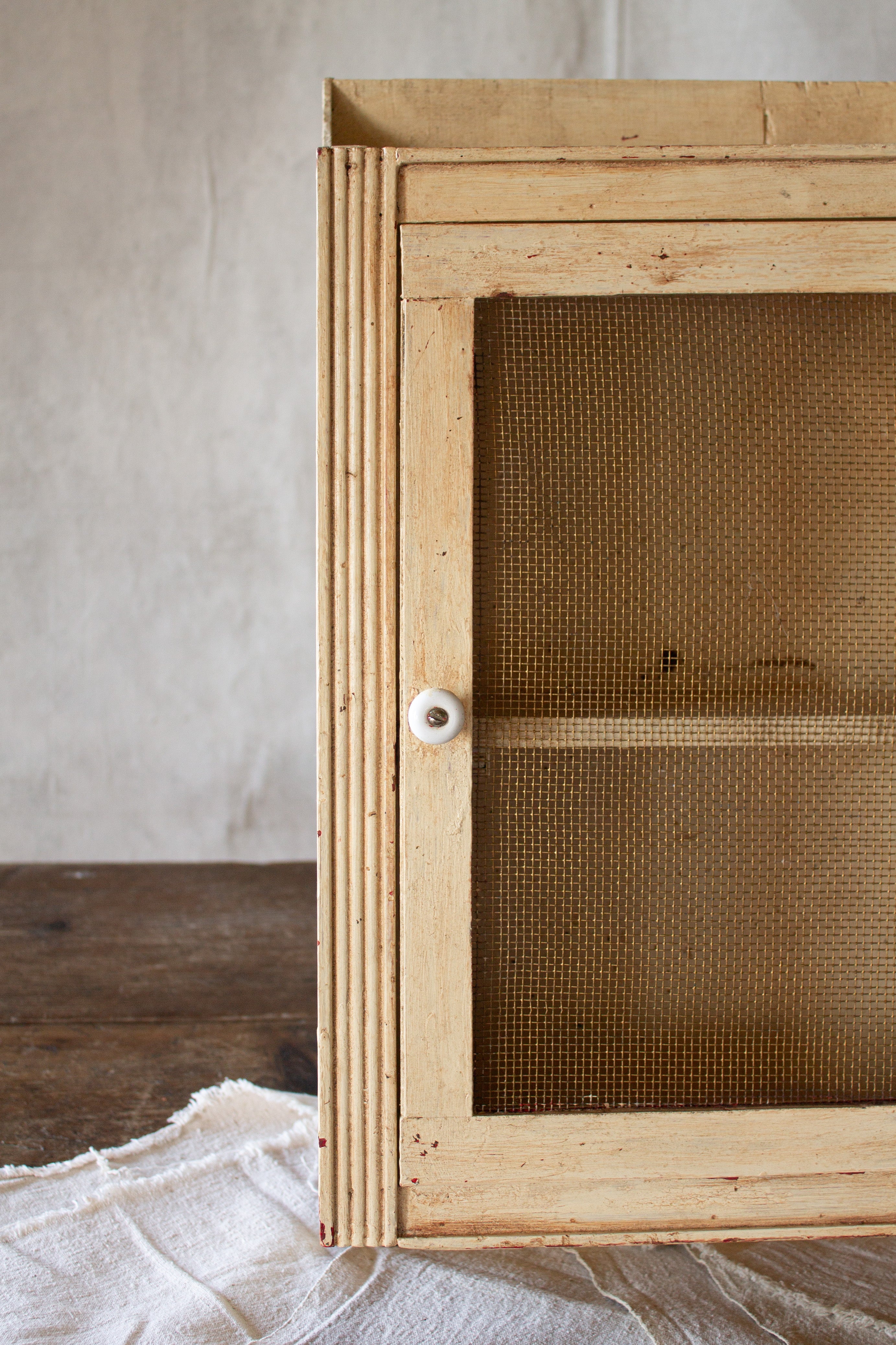 Vintage cream-painted primitive wall cupboard with a wire-mesh door, porcelain knob, fluted trim, and open shelf.