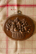 Styled view of vintage copper fruit basket wall mold on antique textile for French country kitchen decor.