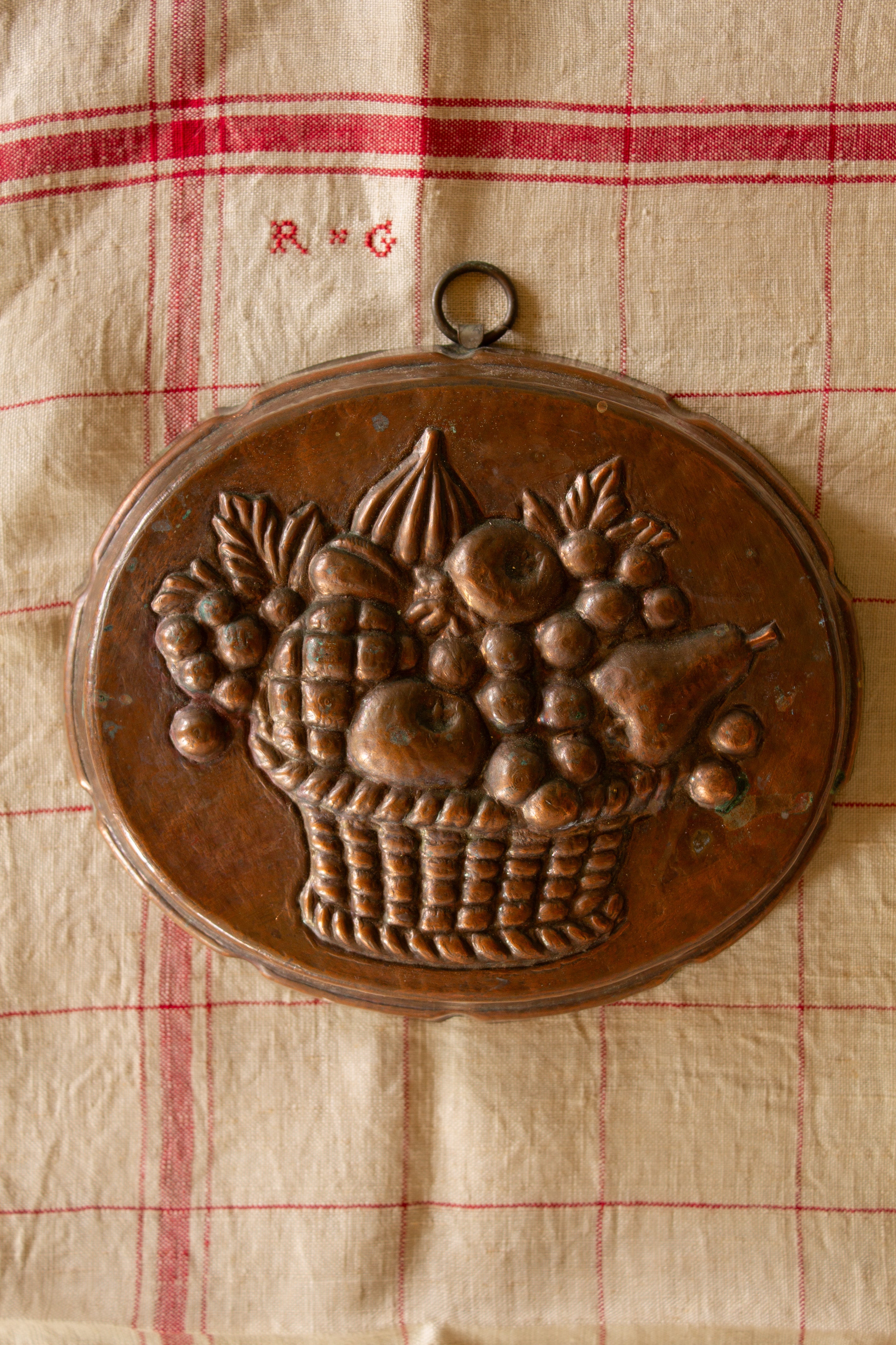 Styled view of vintage copper fruit basket wall mold on antique textile for French country kitchen decor.